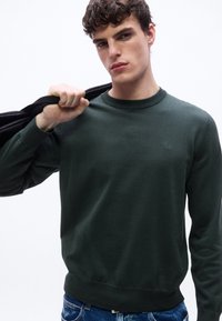 Νεαρός άνδρας που φοράει σκούρο πράσινο πουλόβερ BOSS κρατά ένα μαύρο σακάκι στον ώμο του μπροστά σε απλό λευκό φόντο.