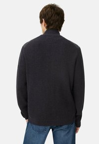 Donkergrijze ribgebreide trui met een hoge kraag. Beschikt over textuur op de schouders en een relaxte pasvorm. Gecombineerd met lichtblauwe jeans.