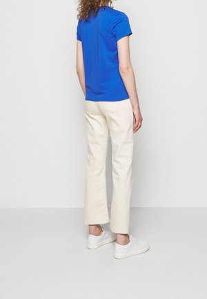 Persoon staat met de rug naar de kijker, draagt een blauw shirt met korte mouwen, crèmekleurige broek en witte sneakers tegen een effen achtergrond.