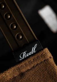 Brown textured fabric strap with a black label featuring white cursive text. Metallic slider with rounded bumps attached at the top.