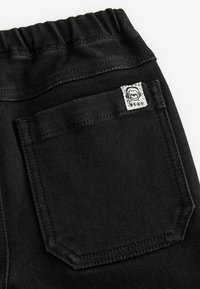 Pantalons en denim noir avec une taille élastique, une poche plaquée à l'arrière et des détails de couture subtils. Étiquette visible sur la poche.