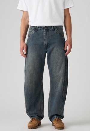 Mann trägt locker sitzende, gewaschene dunkelblaue Jeans mit braunen Schnürschuhen und einem schlichten weißen T-Shirt vor hellem Hintergrund.