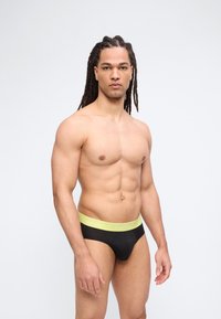 Močan moški z dolgimi spetimi lasmi, oblečen v črne spodnjice z rumenim pasom, stoji ob preprostem svetlem ozadju.