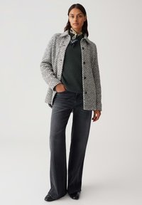 Frau trägt einen schwarzen strukturierten Pullover, eine graue gemusterte Jacke mit Knöpfen, schwarze weite Jeans, schwarze Schuhe und einen grünen gemusterten Hals Schal.