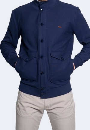 Mann trägt eine dunkelblaue Jacke mit Knöpfen, Klappentaschen und kleinem Hunde-Logo auf der Brust, kombiniert mit beigen Hosen, Hände in den Taschen.