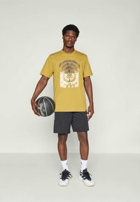 Gul grafisk t-skjorte med et sentralt design i dempede farger, parret med svarte shorts og basketballsko, holder en basketball.