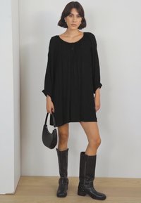 Schwarzes, übergroßes Kleid mit rundem Ausschnitt und langen Ärmeln. Das Model trägt kniehohe schwarze Stiefel und hält eine kleine schwarze Handtasche.