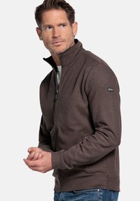 Heren zip-up jas in donkerbruin textuurstof, met een hoge kraag en voorzakken, gestyled met een aansluitende silhoeët.