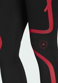 Czarne legginsy o gładkiej teksturze z czerwonymi, zakrzywionymi akcentami oraz detalem z logo na kolanie. Materiał jest elastyczny i dopasowujący się do sylwetki.