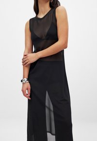 Mustan, hihattoman maksimekon, joka on valmistettu läpinäkyvästä kankaasta ja siinä on sivuhalkio sekä pyöreä pääntie. Mekossa on kerroksellinen muotoilu, jossa alusvaatteet näkyvät.