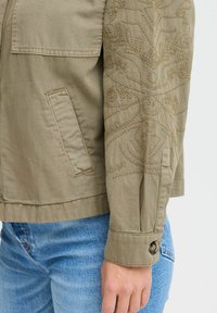 Khaki bunda s vyšitými detailmi na rukáve, so zapínaním na gombík a bočnou kapsou. V súprave s modrými džínsami.