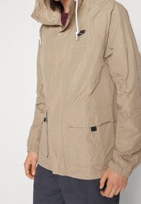 Beige lätt jacka med en texturerad yta, huva, framfickor och elastiska manschetter. Har en liten svart Nike-logotyp på bröstet.