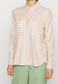 Camicia a righe con bottoni in morbido bianco e pesche, dotata di due tasche sul petto e un orlo leggermente curvo. Realizzata in tessuto leggero.