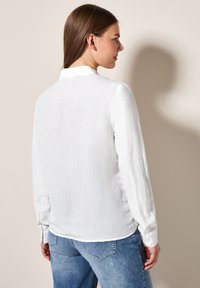 Camicia bianca a maniche lunghe realizzata in tessuto leggero, con colletto, polsini abbottonati e taglio ampio, abbinata a jeans blu.