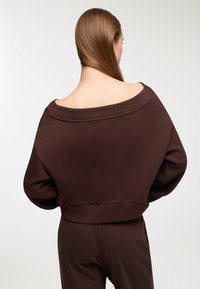 Felpa marrone scesa sulle spalle con vestibilità ampia, scollatura larga e orlo a coste. Il tessuto sembra morbido con una texture liscia.