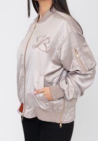 Femme portant une veste bomber en satin beige clair avec la lettre B brodée, fermeture éclair devant, poignets côtelés et poche zippée sur la manche gauche.