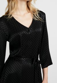 Abito nero con scollo a V, caratterizzato da un motivo a diamante testurizzato, maniche lunghe, chiusura con bottoni e cintura annodata in vita.