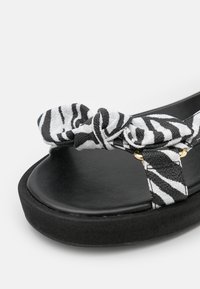 Sandale rayée zèbre en noir et blanc avec une bride en tissu nouée et une semelle plateforme noire lisse ornée de détails métalliques dorés.