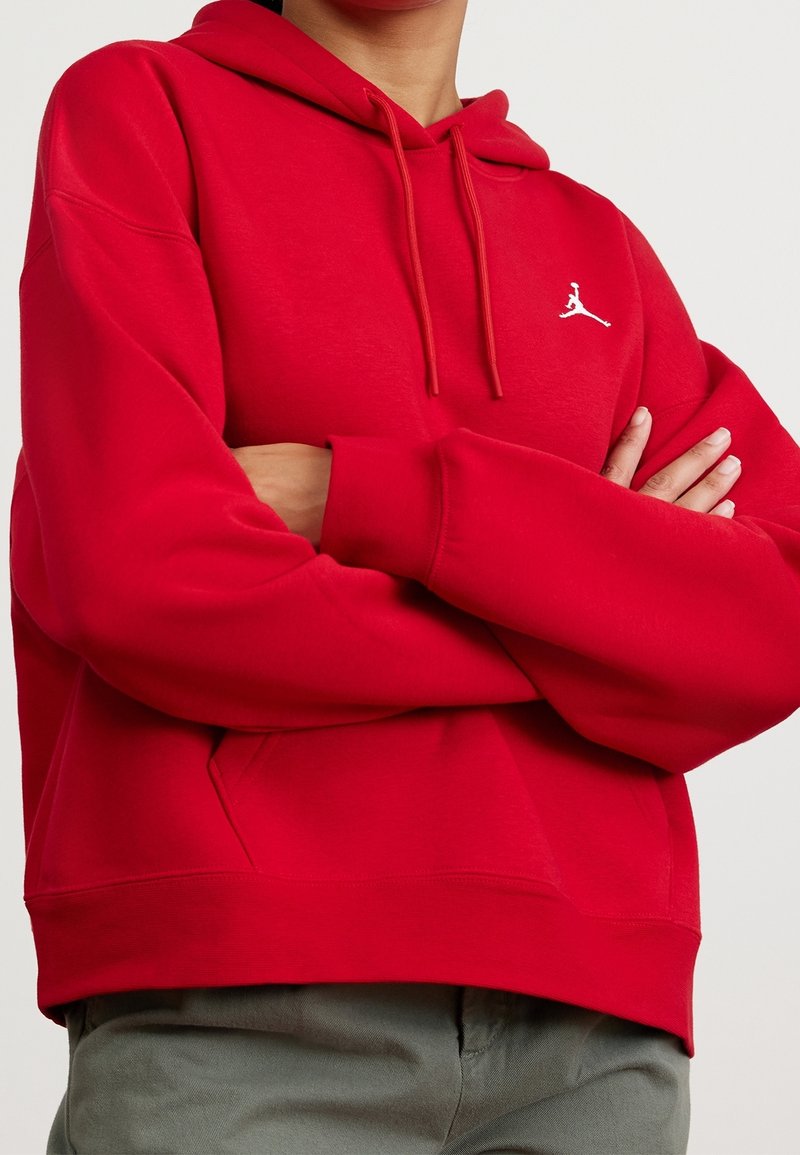 Personne portant un sweat à capuche rouge, les bras croisés, avec un petit logo blanc Jumpman sur la poitrine, associé à un pantalon gris.