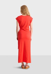 Robe maxi rouge en tissu doux, avec des manches courtes et un ajustement décontracté, dotée d'une fente sur le côté. Portée avec des sandales à talons bruns.