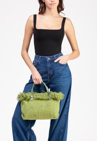 Borsa a tracolla intrecciata verde con cima frangiata, accessori metallici e testo ricamato. Il modello indossa una maglietta nera e jeans a vita alta blu.