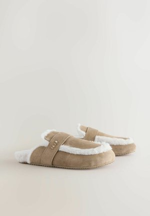 Tanfarbene Wildlederschuhe mit weißem Futter und Innenauskleidung. Verfügt über einen Riemen mit goldener Schrift. Weiche Textur und flache Sohle.