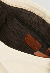 Interior de bolso de lona, tela color crema con un bolsillo con cremallera. El forro a cuadros marrón oscuro presenta una etiqueta de cuero. Diseño suave y estructurado.