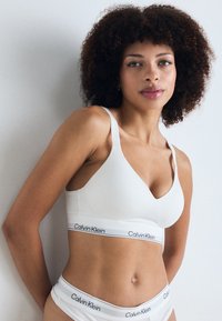 Witte Calvin Klein bralette met een v-halsontwerp, zachte stof en een brede band met het merklogo. Witte low-rise slipjes zijn ook zichtbaar.