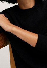 Pull en tricot noir avec un col rond et des poignets côtelés, mettant en valeur une texture lisse et un design ajusté, posé sur une surface en bois.