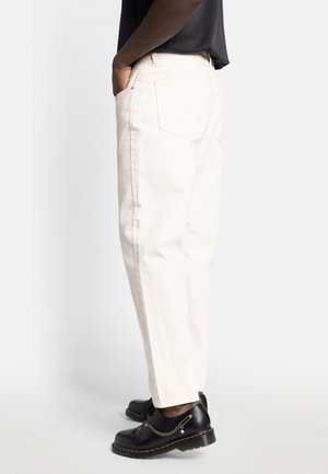 Personne portant un pantalon droit couleur blanc cassé, une chemise noire et des chaussures en cuir noir avec un détail en chaîne métallique, debout devant un fond blanc.