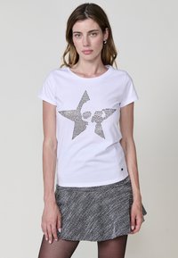 Camiseta de algodón blanca con un diseño de estrella negra hecho de pequeños tachuelas. Cuello redondo y mangas cortas. Combinada con una falda negra texturizada.