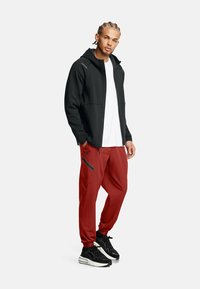 Schwarze Zip-Up-Hoodie mit glatter Textur, kombiniert mit roten Jogginghosen mit seitlichen Reißverschlusstaschen und schwarzen Sneakers mit weißen Akzenten.