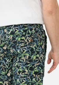 Dunkelblaue Shorts mit floralen Mustern in grünen und beigen Blättern. Mit einer Seitentasche und sichtbaren Nähdetails.