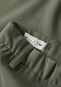 Maslinasto zelena tkanina s glatkom teksturom, koja sadrži bijelu oznaku i jednim dugmetom za zatvaranje, s naboranim detaljima uz rub.