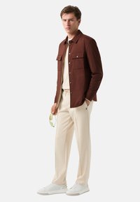Braune leichte Jacke mit zwei vorderen Taschen, beigem Oberteil, cremefarbener Hose und weißen Sneakers. Das Modell hält grüne Sonnenbrillen.