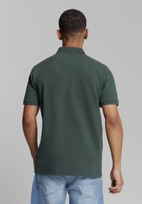 Donkergroen polo met korte mouwen en een opstaande kraag, met een gladde textuur en een rechte zoom. Draagt een lichte spijkerbroek.
