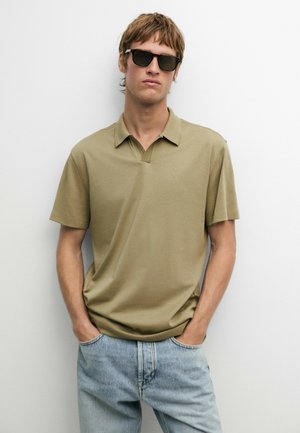 Giovane uomo con occhiali da sole scuri, polo beige a maniche corte e jeans azzurri, in piedi davanti a un muro bianco semplice.