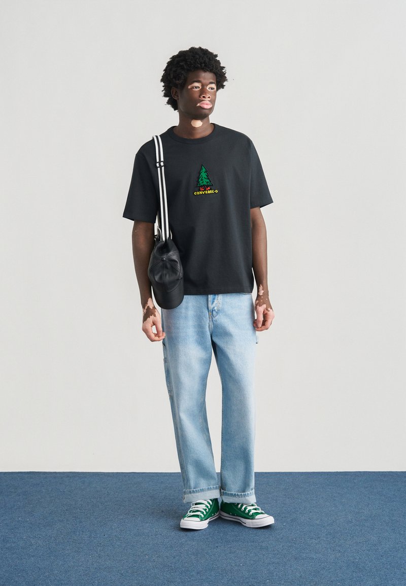 Maglietta nera a maniche corte con logo di un albero verde e testo, jeans larghi azzurro chiaro, sneakers Converse verdi e borsa a tracolla nera.