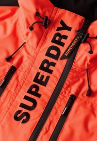 Veste orange vif avec des fermetures éclair noires, des cordons et un grand logo noir "SUPERDRY" brodé sur la zone de la fermeture éclair avant.