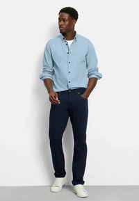 Hellblaues Hemd mit Knöpfen, dunkle Jeans und weiße Turnschuhe. Das Hemd hat eine lockere Passform und hochgekrempelte Ärmel. Das Modell steht mit den Händen in den Taschen.