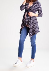 Mörkblå omslagströja med geometriskt mönster, långa ärmar och knytning, matchad med blå skinny jeans och vita sneakers.