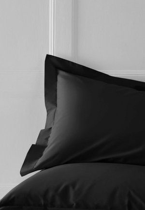 Schwarzes Kissen mit breitem Rand auf einem passenden schwarzen Bett vor einer weißen, vertäfelten Wand.
