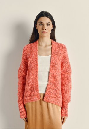 Helle korallenfarbene Strickjacke mit lockerer Passform, weicher Textur, offenem Vorderteil und gerippten Ärmeln. Getragen über einem weißen Oberteil und einem beigen Rock.