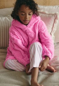 Couverture à capuche rose flou avec une surface texturée ; associée à un pantalon de pyjama rayé rose et blanc. Assise sur une couverture tissée.