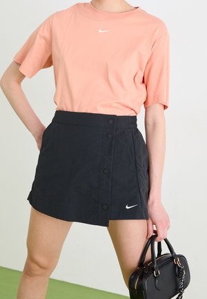 Osoba ubrana w brzoskwiniową koszulkę Nike i czarną spódnicę Nike, trzymająca czarną skórzaną torebkę z złotymi detalami zamka błyskawicznego.