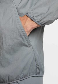 Giacca leggera grigia con polsini elastici e tasca laterale, caratterizzata da una texture liscia e leggermente stropicciata e da un design minimalista.