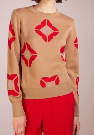 Personne portant un pull couleur chameau avec des motifs géométriques rouges en forme de losange et un pantalon rouge, montré du cou aux hanches.
