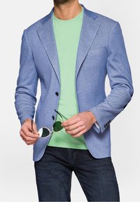 Světle modrý strukturovaný blazer se dvěma černými knoflíky, nošený přes mátově zelené tričko. Model drží sluneční brýle se zeleným nádechem. Tmavé džíny dotvářejí celkový outfit.