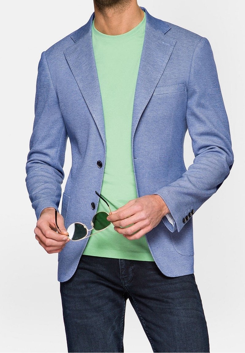 Světle modrý strukturovaný blazer se dvěma černými knoflíky, nošený přes mátově zelené tričko. Model drží sluneční brýle se zeleným nádechem. Tmavé džíny dotvářejí celkový outfit.