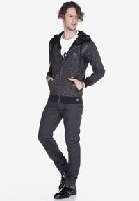Fleece-Zip-Hoodie in Dunkelgrau mit schwarzen Akzenten, versehen mit einem strukturierten Design, einer Kängurutasche und einer glatten Kapuze. Schwarze, eng anliegende Hose und glänzende Stiefel.